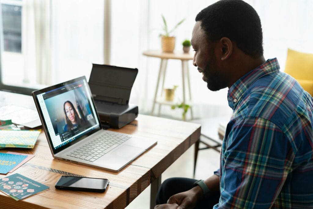 A man sits at a wooden desk, participating in a video call on a laptop. Printed papers, a smartphone, and a printer are on the desk. A woman is visible on the laptop screen.