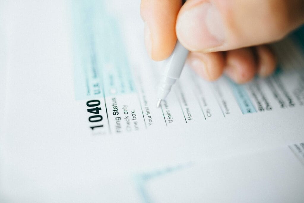 A close-up of a hand holding a pen, filling out a U.S. IRS 1040 tax form. The focus is on the top of the tax form and the pen, with some text visible but not fully legible.