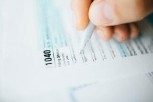 A close-up of a hand holding a pen, filling out a U.S. IRS 1040 tax form. The focus is on the top of the tax form and the pen, with some text visible but not fully legible.