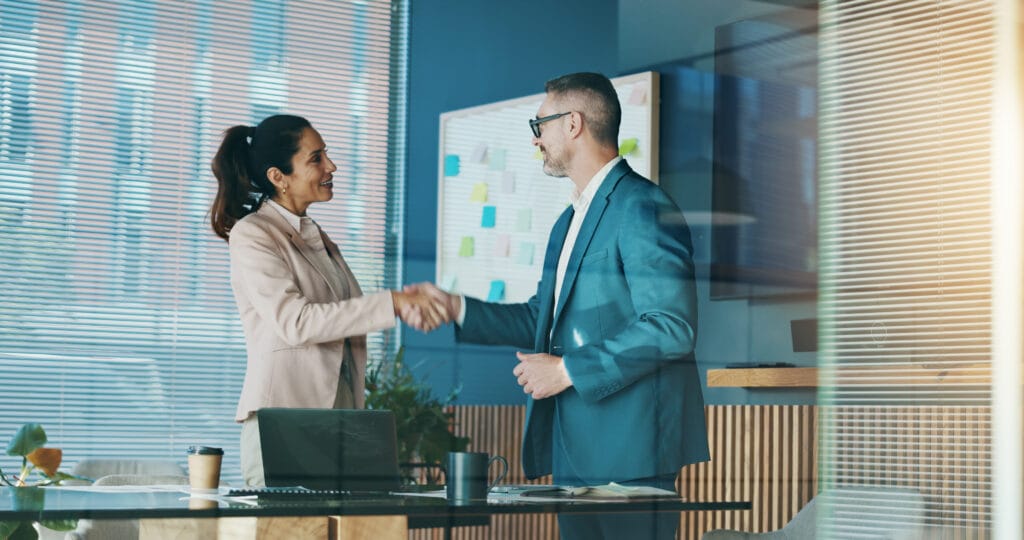 A woman and a man in business attire shake hands in a modern office. A laptop, cup, and papers are on the desk. Blinds cover the windows, and a whiteboard with sticky notes is visible in the background.