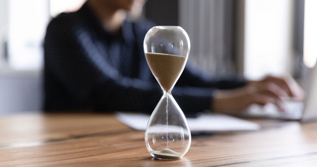 A glass hourglass with sand flowing through it is on a wooden table. In the background, a person is typing on a laptop, partially out of focus.