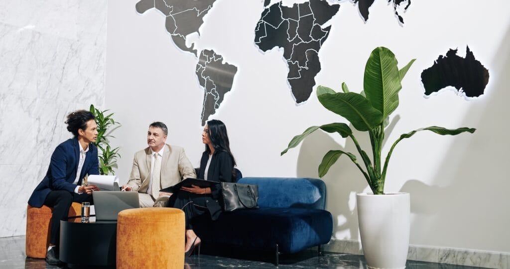 Three people in business attire sit and talk in a modern office lounge with a blue sofa, yellow ottoman, potted plants, and a large world map on the wall behind them.