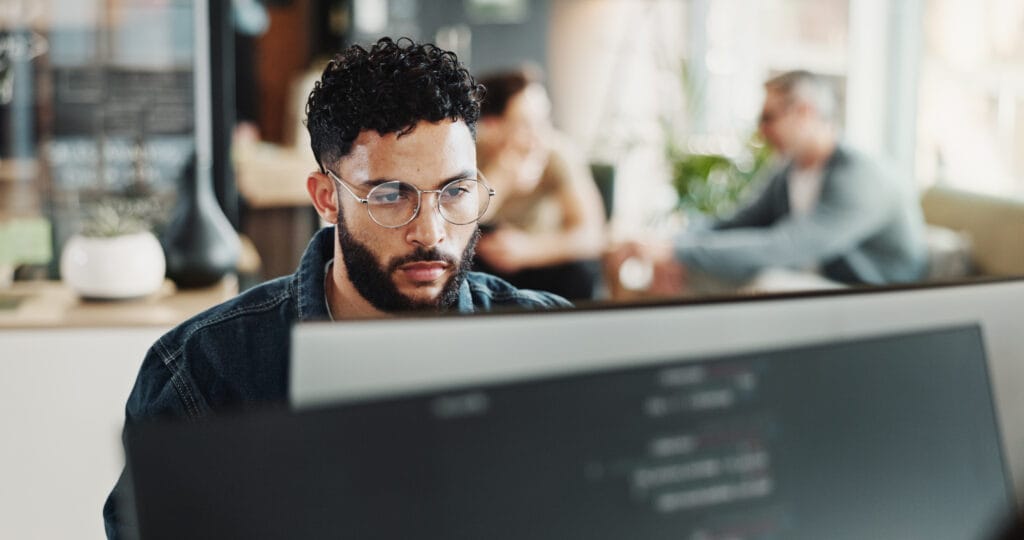 A man with glasses and curly hair focuses on a computer monitor in an office setting, with other people sitting and talking in the blurred background.