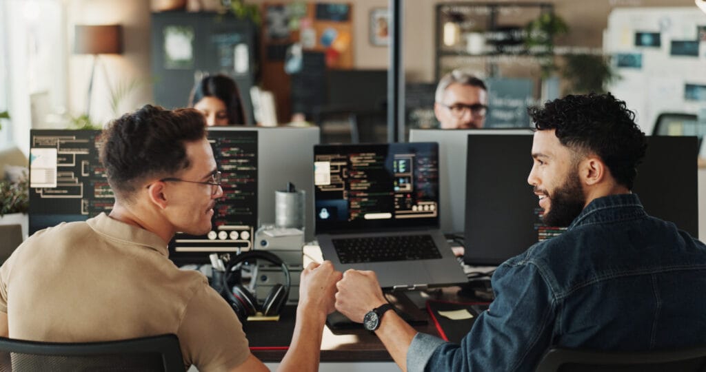 Two men sitting at a desk in an office environment, giving each other a fist bump. Computer monitors with code are visible on the desks, and other people are working in the background.