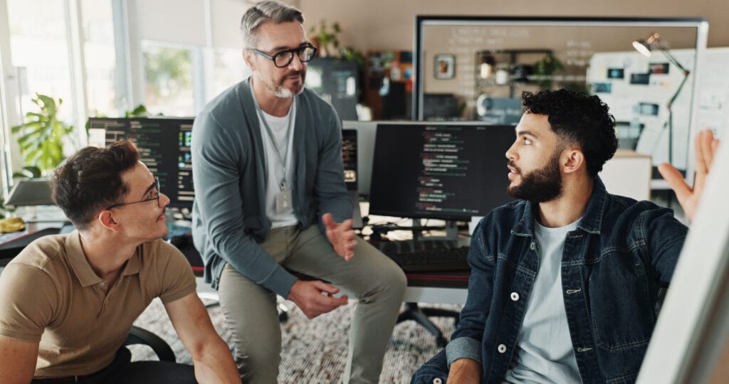 Three men sit and talk in an office with computer monitors displaying code in the background. One man gestures with his hand while the others listen, appearing engaged in conversation.