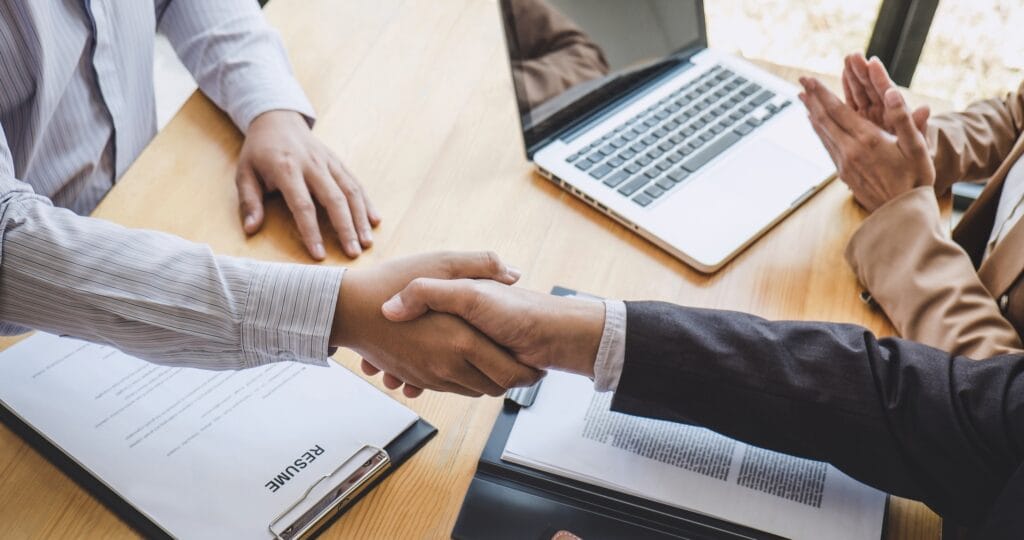 Two people are shaking hands across a desk with resumes, documents, and a laptop. A third person sits nearby with hands clasped, suggesting a professional meeting or job interview.