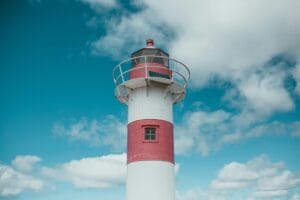 A white lighthouse with a red horizontal stripe and balcony stands against a blue sky with scattered white clouds.