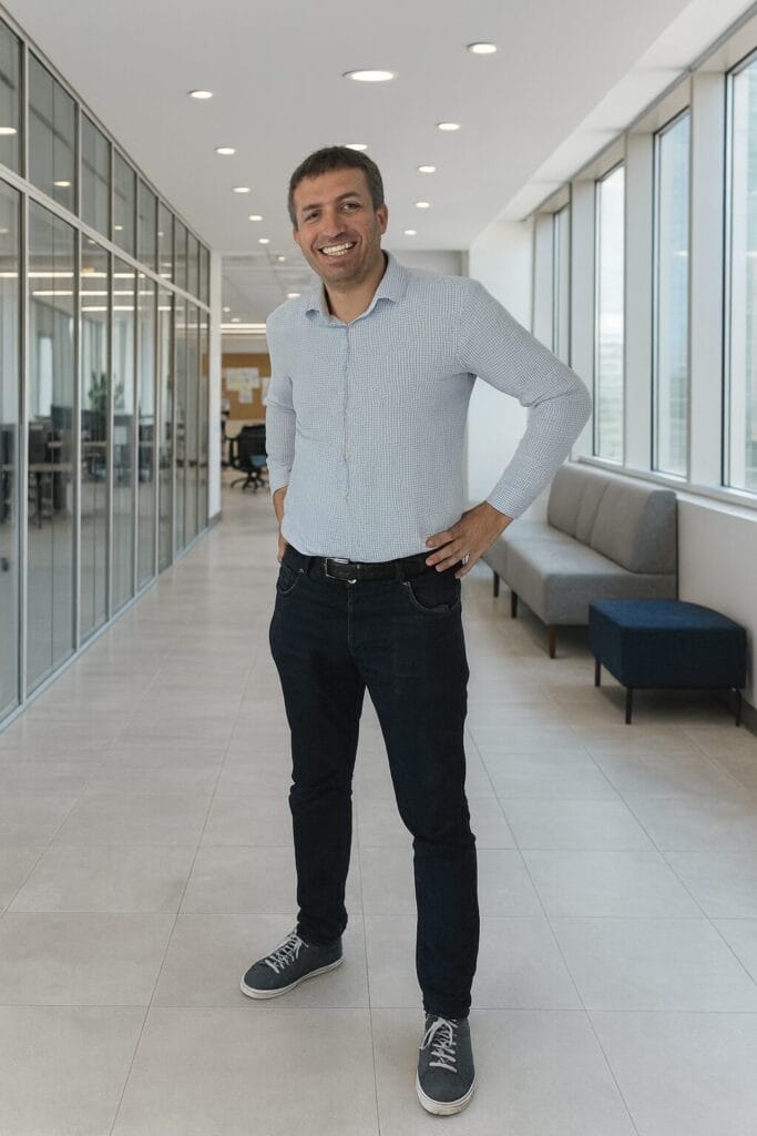 A man stands in a bright, modern office hallway with large windows, hands on his hips, smiling at the camera. He is wearing a light-colored shirt, dark jeans, and sneakers.